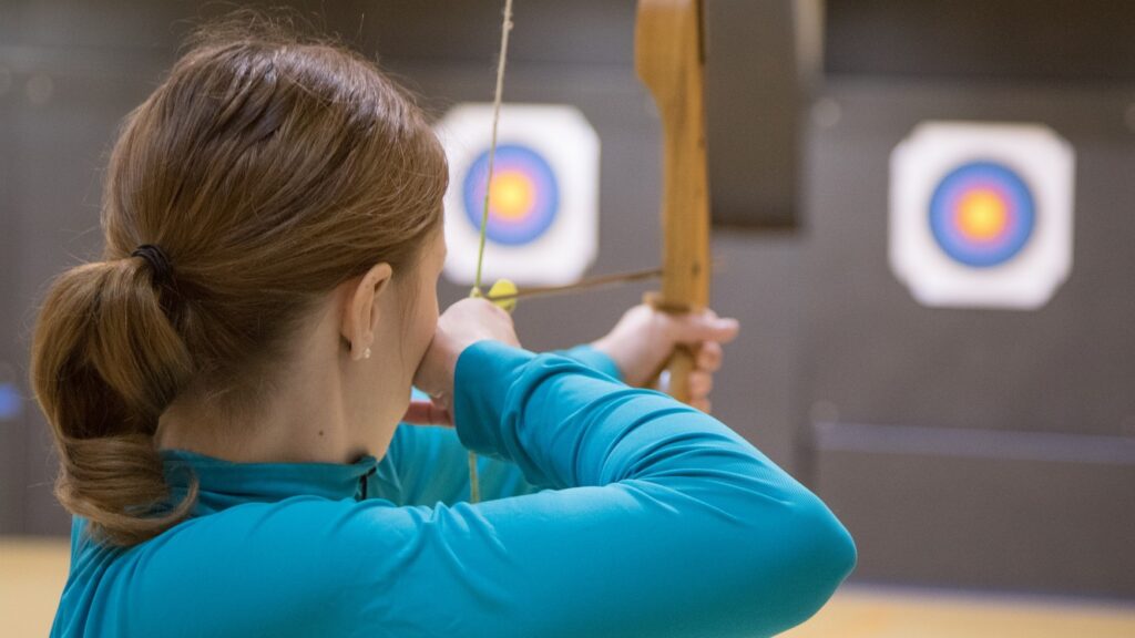 A woman takes aim with a bow and arrow at a target in the distance.