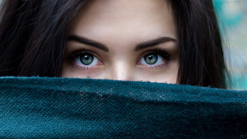 A girl with black hair and green eyes gazes into the camera. the lower half of her face is obscured.