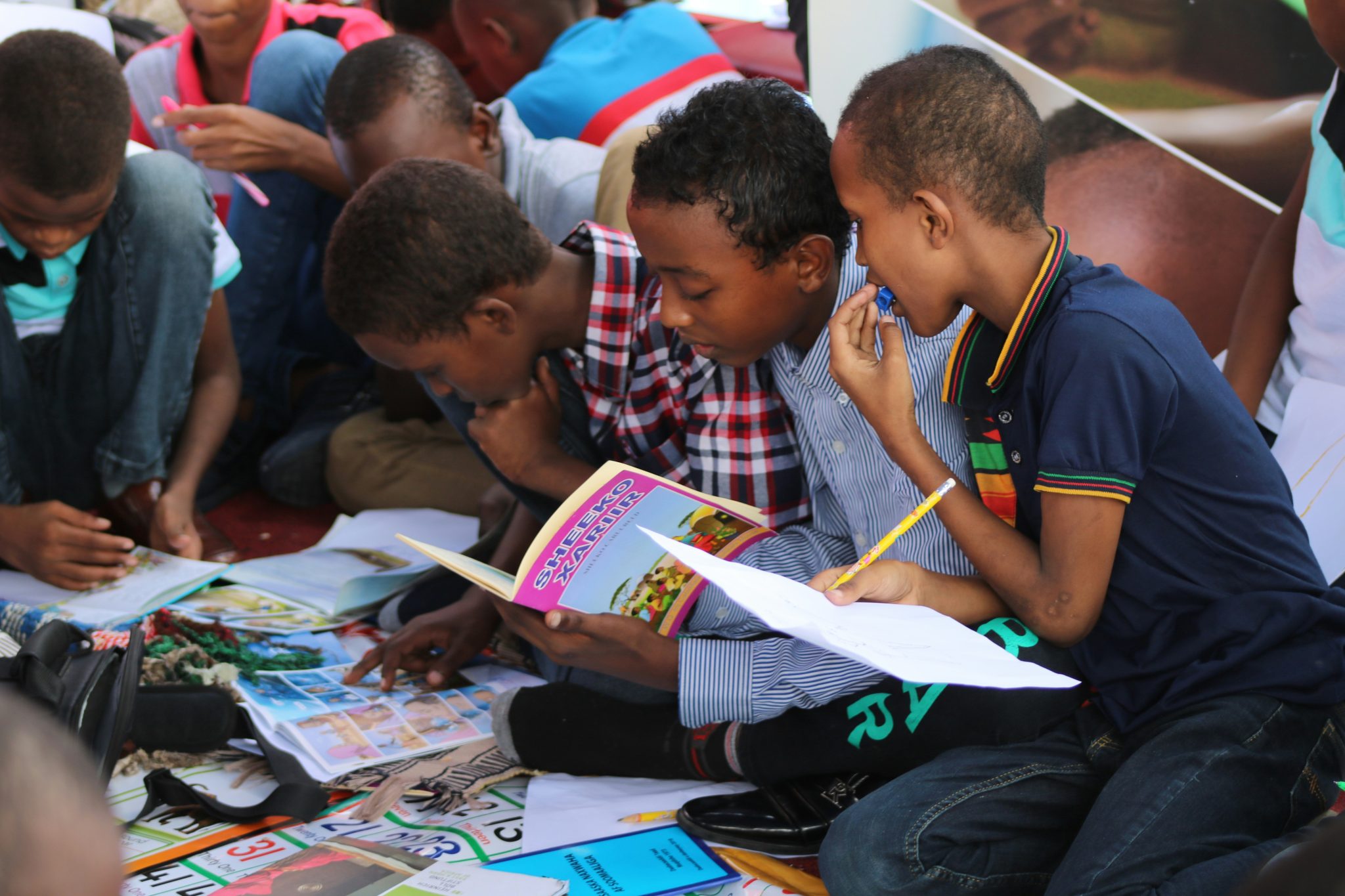 Three boys sit on the floor of a classroom, absorbed in the books they are reading. All three boys are black; one is wearing a plaid button-down shirt, the second has on a blue and white-striped button-down shirt, and the third wears a blue polo.